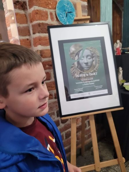 hdimg01c39240647ea185ee08cecaddcdd0 chłopak w niebieskiej kurtce, za nim plakat "Twarzą w twarz" na sztaludze